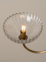 A close-up of a wall-mounted light fixture with a clear ribbed glass shade in a scalloped rim design, mounted on a brushed brass metal arm against a warm beige background.