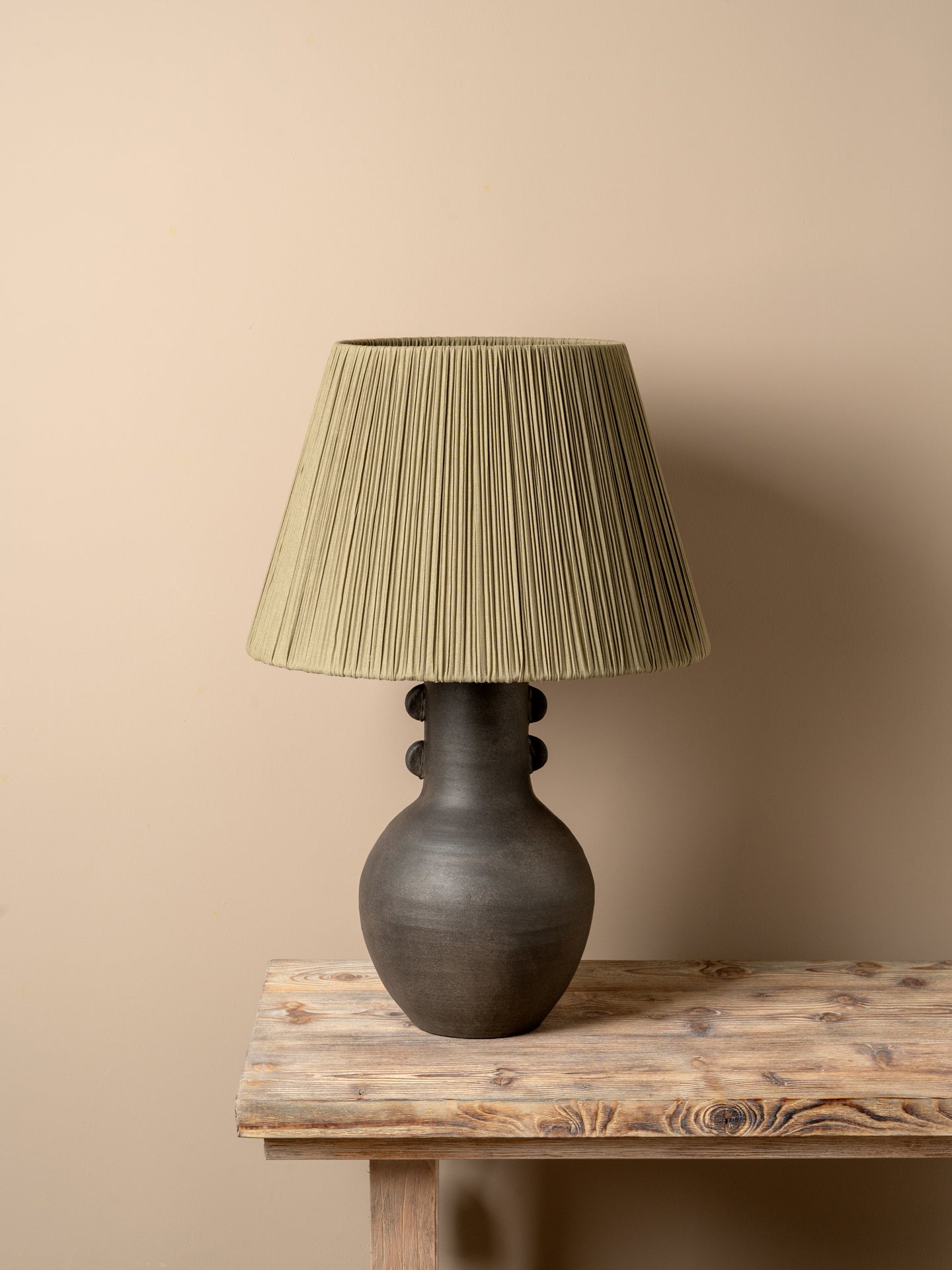 A table lamp with a dark brown ceramic vase-shaped base and a pleated olive green fabric shade, illuminated with a warm glow on a rustic wooden table against a neutral beige wall.