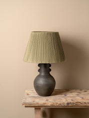 A table lamp with a dark brown ceramic vase-shaped base and a pleated olive green fabric shade, illuminated with a warm glow on a rustic wooden table against a neutral beige wall.