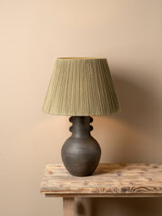 A table lamp with a dark brown ceramic vase-shaped base and a pleated olive green fabric shade, illuminated with a warm glow on a rustic wooden table against a neutral beige wall.
