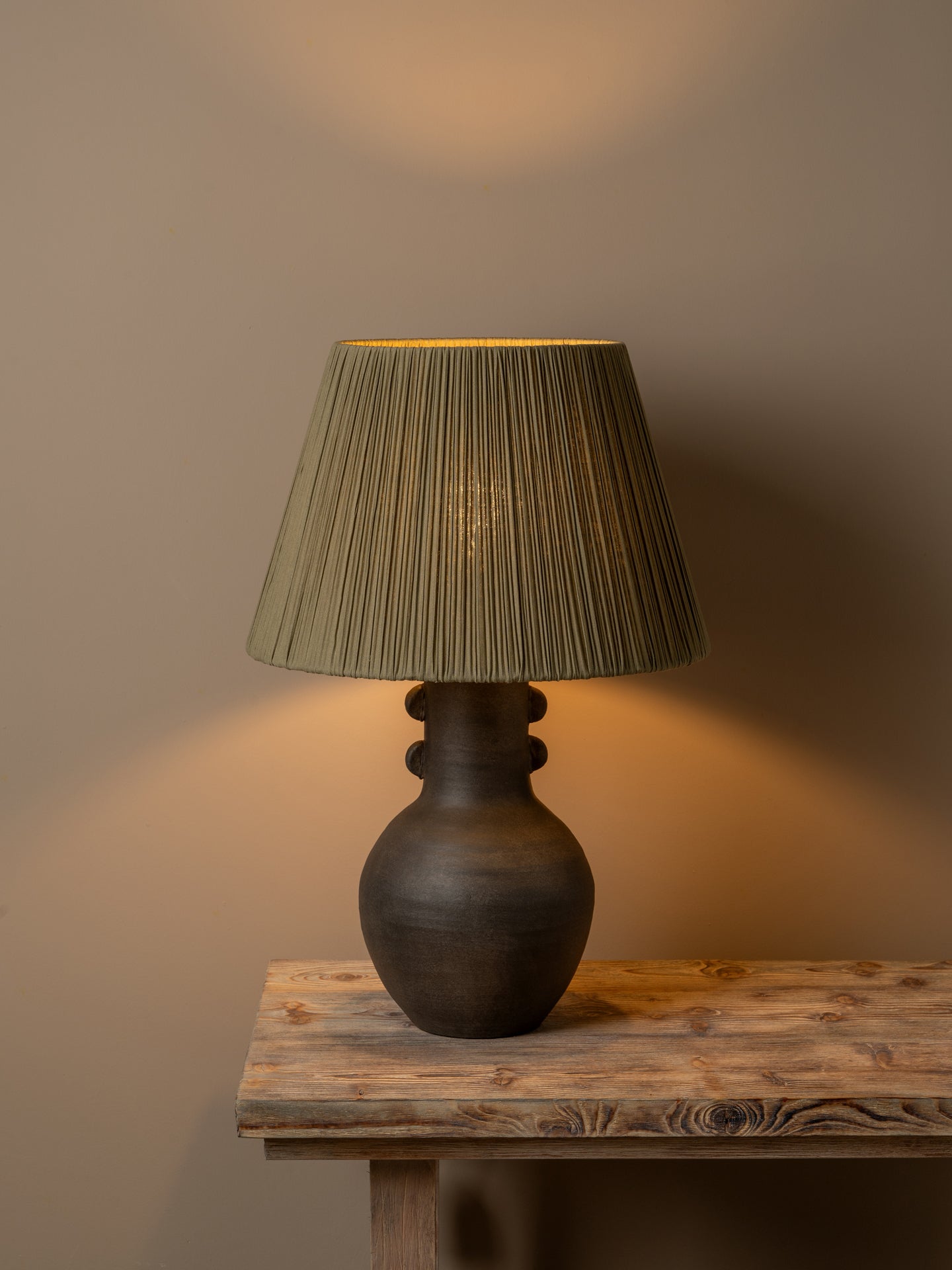 A table lamp with a dark brown ceramic vase-shaped base and a pleated olive green fabric shade, illuminated with a warm glow on a rustic wooden table against a neutral beige wall.