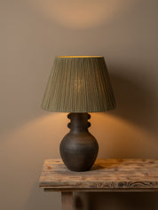 A table lamp with a dark brown ceramic vase-shaped base and a pleated olive green fabric shade, illuminated with a warm glow on a rustic wooden table against a neutral beige wall.