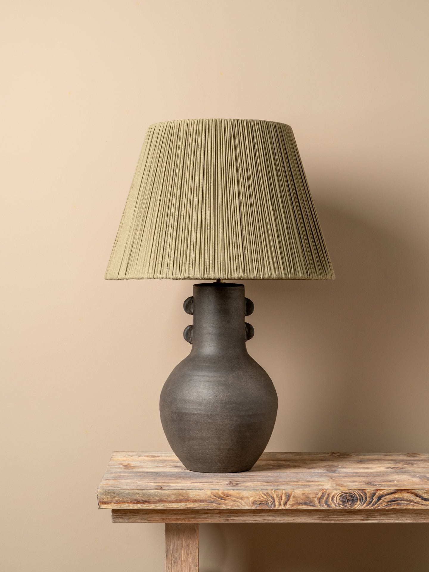 A table lamp with a dark brown ceramic vase-shaped base and a pleated olive green fabric shade, illuminated with a warm glow on a rustic wooden table against a neutral beige wall.