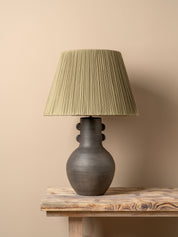A table lamp with a dark brown ceramic vase-shaped base and a pleated olive green fabric shade, illuminated with a warm glow on a rustic wooden table against a neutral beige wall.