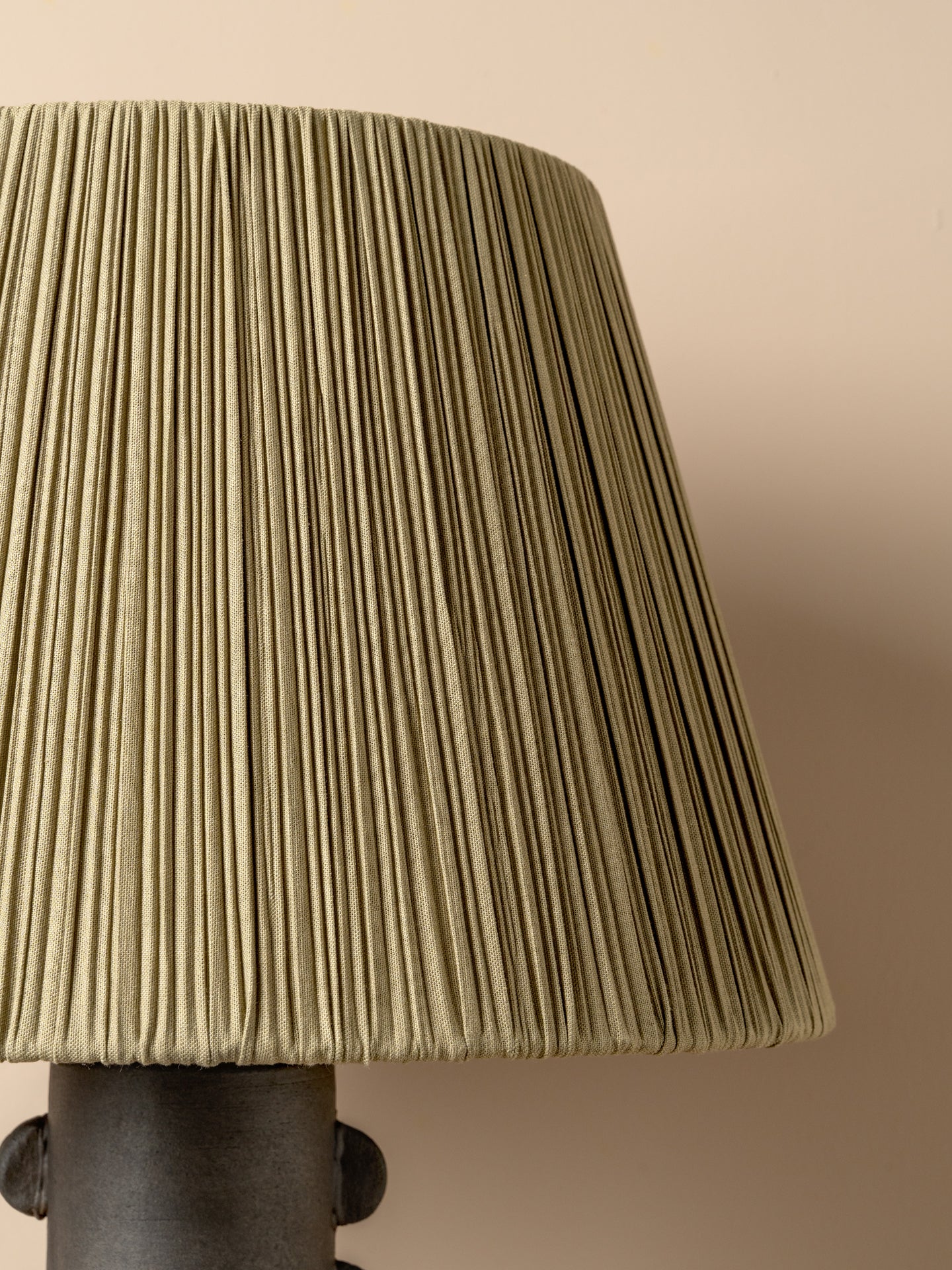 A close-up of a pleated fabric lampshade in a muted olive green color, attached to a dark metal lamp base against a solid beige background.