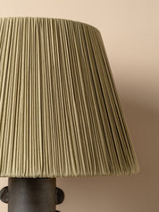 A close-up of a pleated fabric lampshade in a muted olive green color, attached to a dark metal lamp base against a solid beige background.