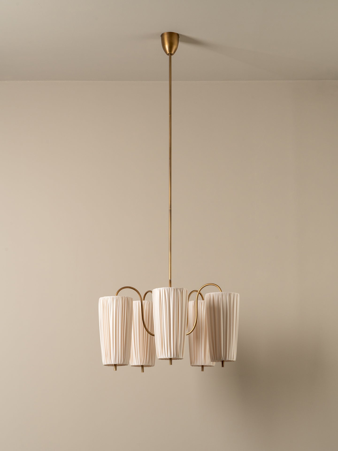 A modern multi-light pendant chandelier with pleated off-white cylindrical shades and a brushed brass finish, suspended from a matching ceiling canopy against a neutral beige wall.