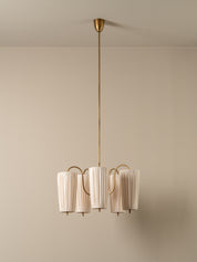 A modern multi-light pendant chandelier with pleated off-white cylindrical shades and a brushed brass finish, suspended from a matching ceiling canopy against a neutral beige wall.