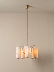 A modern multi-light pendant chandelier with four cylindrical pleated white shades and a brushed brass/gold metal frame and canopy, suspended against a neutral beige wall and ceiling.