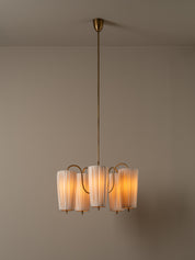 A modern multi-light chandelier with pleated white glass shades and a warm brass/gold metal frame, suspended from a matching ceiling canopy against a neutral beige wall.