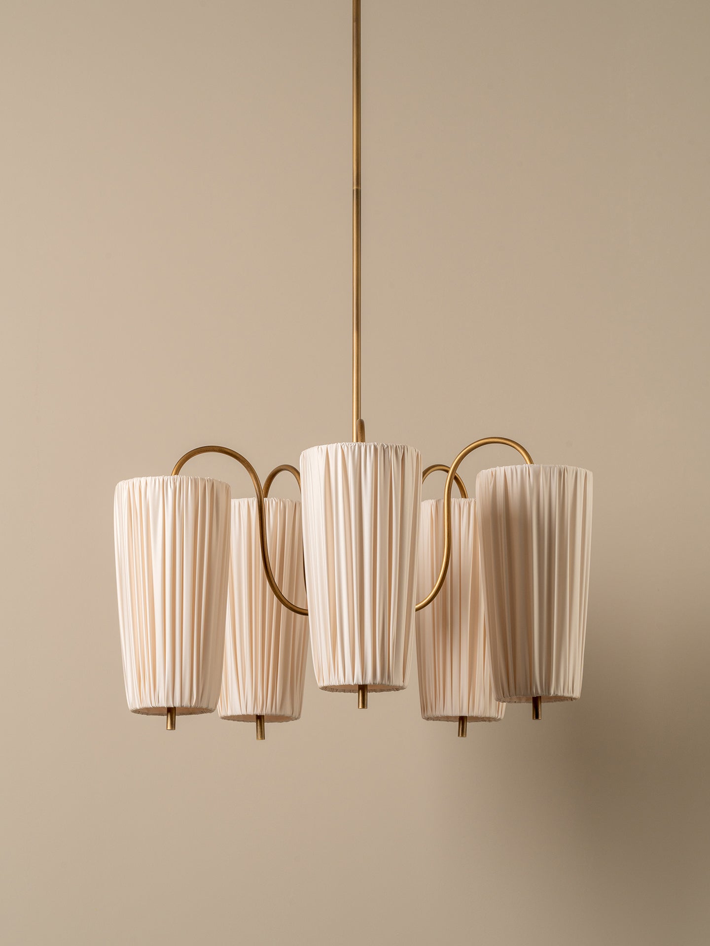 A modern six-light chandelier with pleated off-white fabric shades and a brushed brass metal frame, suspended against a solid beige wall.
