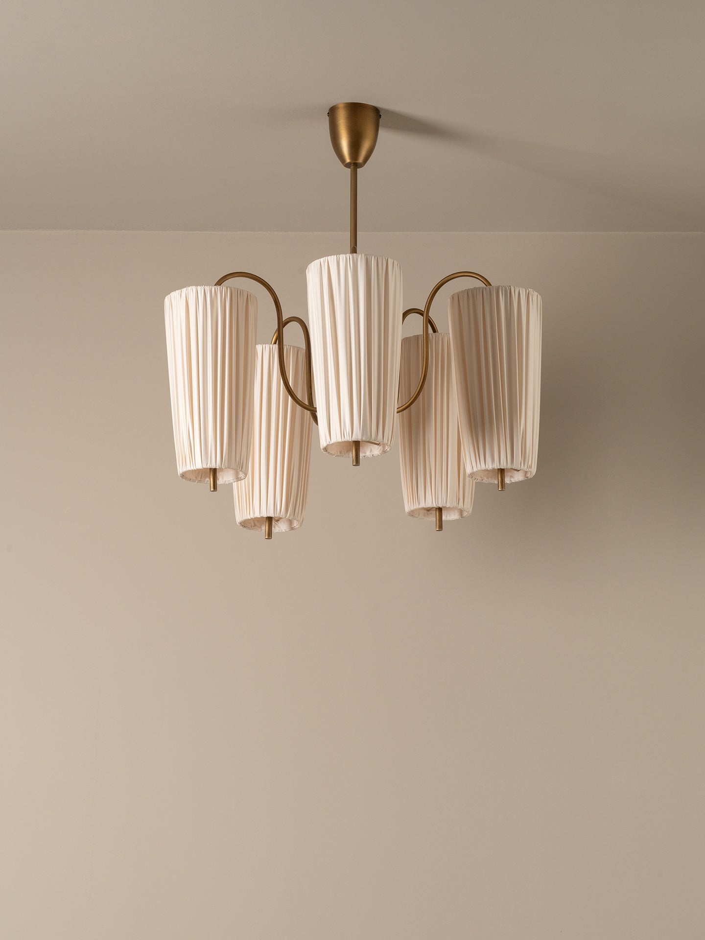A modern five-light chandelier with pleated off-white cylindrical shades and a brushed brass metal frame, suspended from a matching ceiling canopy against a neutral beige wall.