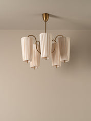A modern five-light chandelier with pleated off-white cylindrical shades and a brushed brass metal frame, suspended from a matching ceiling canopy against a neutral beige wall.