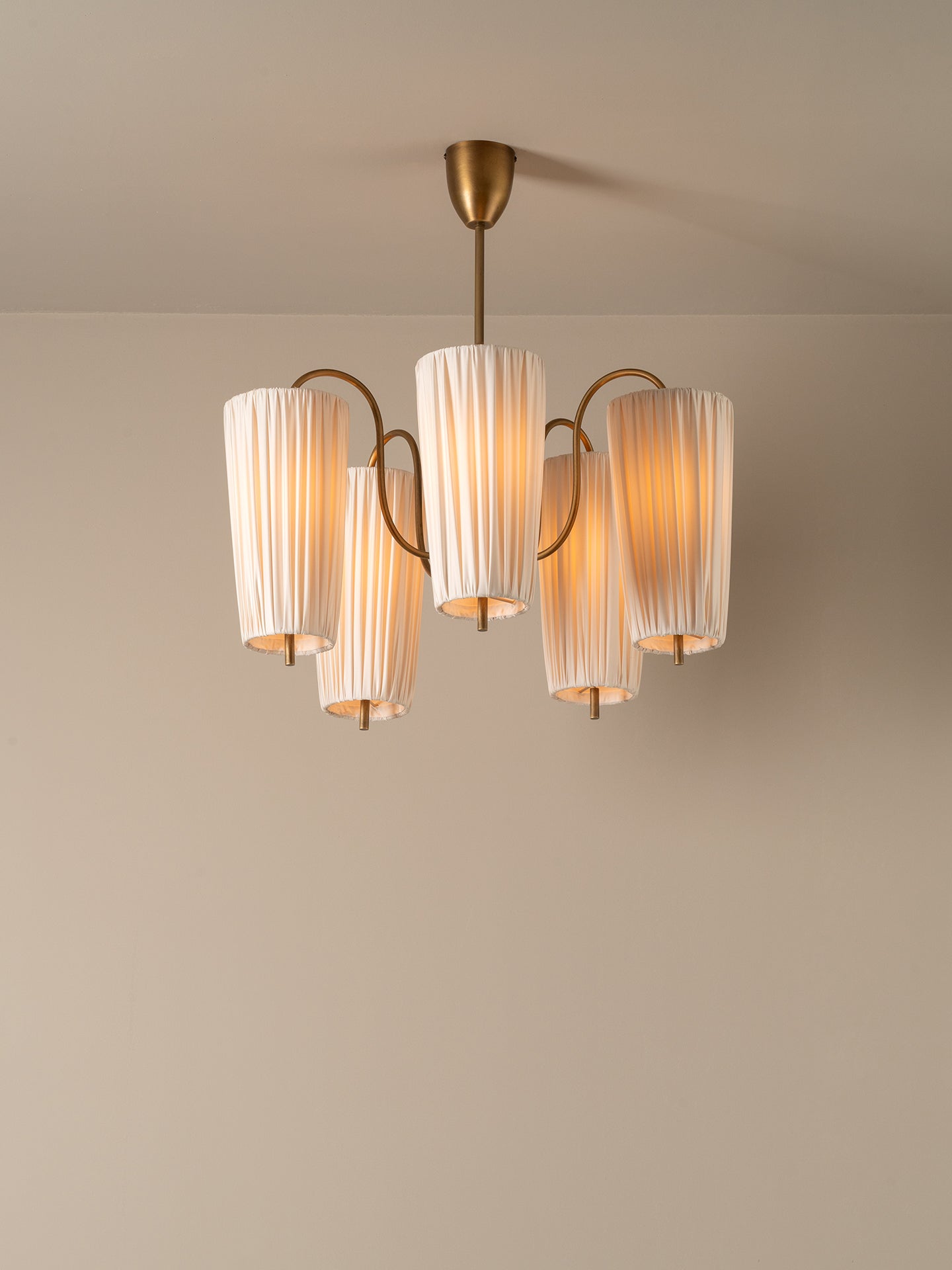 A modern five-light chandelier with pleated white glass shades and a brushed brass finish, suspended from the ceiling against a neutral beige wall.