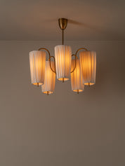 A modern five-light chandelier with pleated white glass shades and a warm brass-finished metal frame, suspended from a ceiling against a neutral taupe wall.
