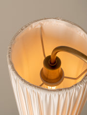 Close-up of a pleated off-white fabric lampshade with a warm yellow glow from an illuminated bulb inside, connected to a brass-colored socket and cord.