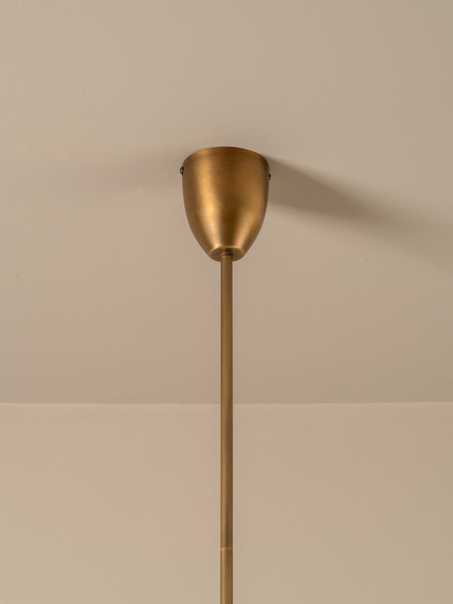 A modern pendant light fixture with a brushed brass or bronze metal shade and a long matching stem, shown hanging against a warm beige wall.