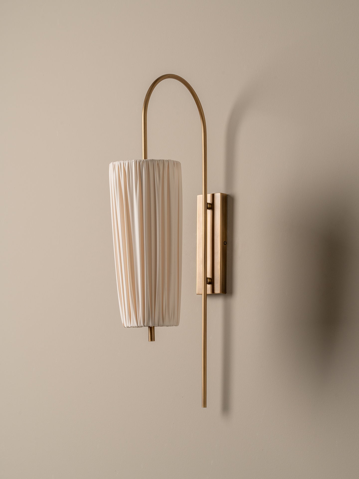 A modern wall-mounted sconce with a vertical pleated white drum shade and a curved brass metal frame, mounted on a beige wall.