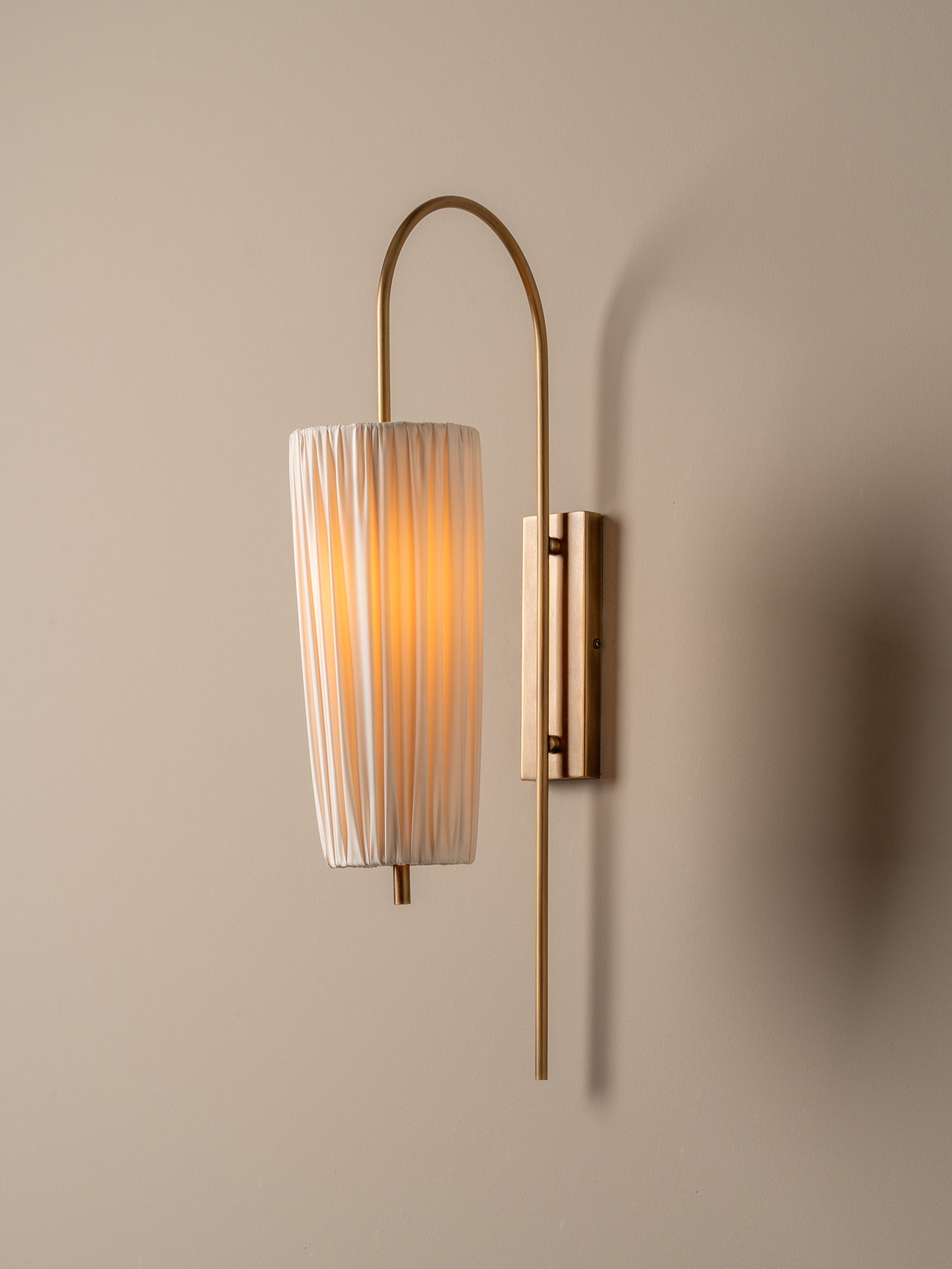 A modern wall sconce with a vertical ribbed white glass shade, a curved gold metal top arm, and a rectangular gold wall mount, shown illuminated against a warm beige wall.