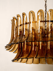 A close-up of a modern chandelier featuring multiple curved, amber-tinted glass shades suspended in a tiered arrangement, with a dark metal chain visible at the top against a plain off-white background.