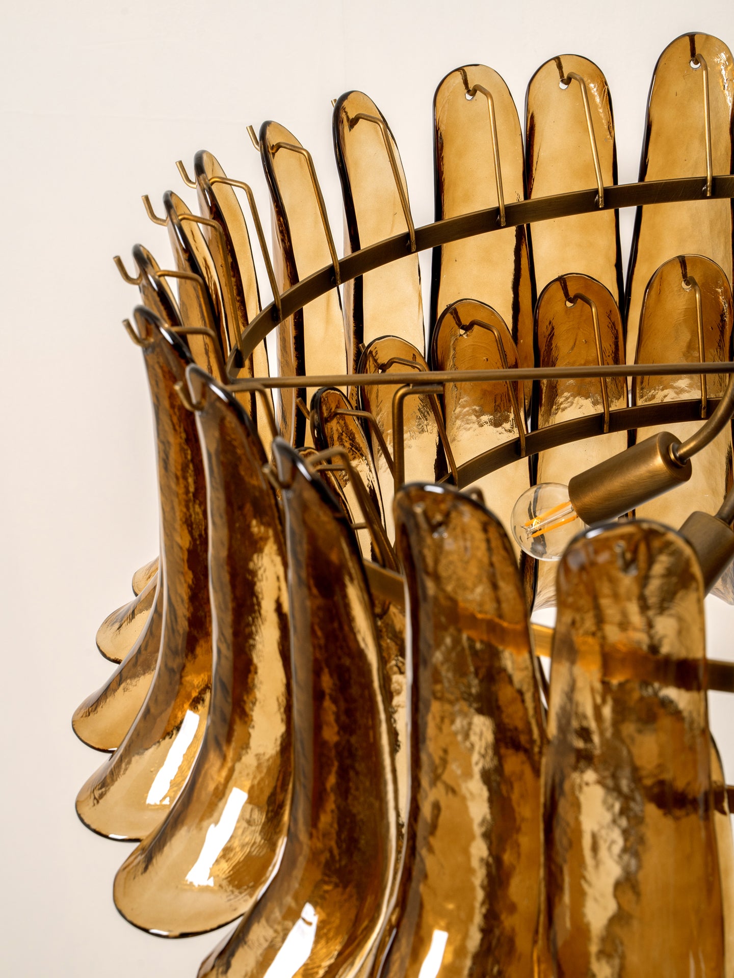 A close-up of a modern chandelier featuring curved, translucent amber-brown glass leaf-like shades suspended in a circular brass metal frame with visible light bulbs.