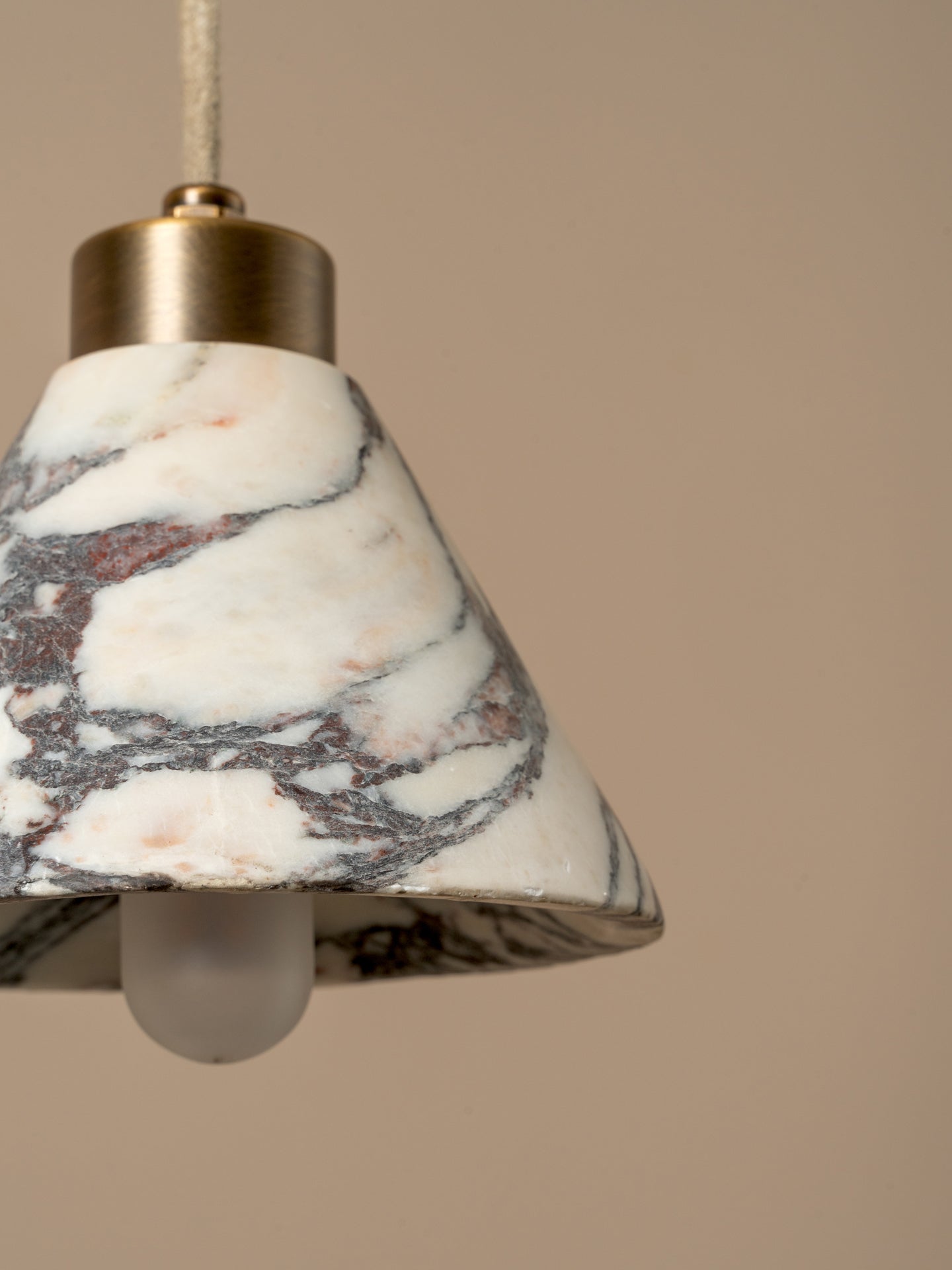 A close-up of a pendant light with a white marble shade featuring gray and subtle pink veining, a brass metal top, and a frosted glass bulb, set against a solid beige background.