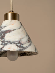 A close-up of a pendant light with a white marble shade featuring gray and subtle pink veining, a brass metal top, and a frosted glass bulb, set against a solid beige background.