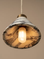 A pendant light with a white marble shade featuring dark brown and gray veining, suspended by a white cord with a brass fitting, against a warm beige background.