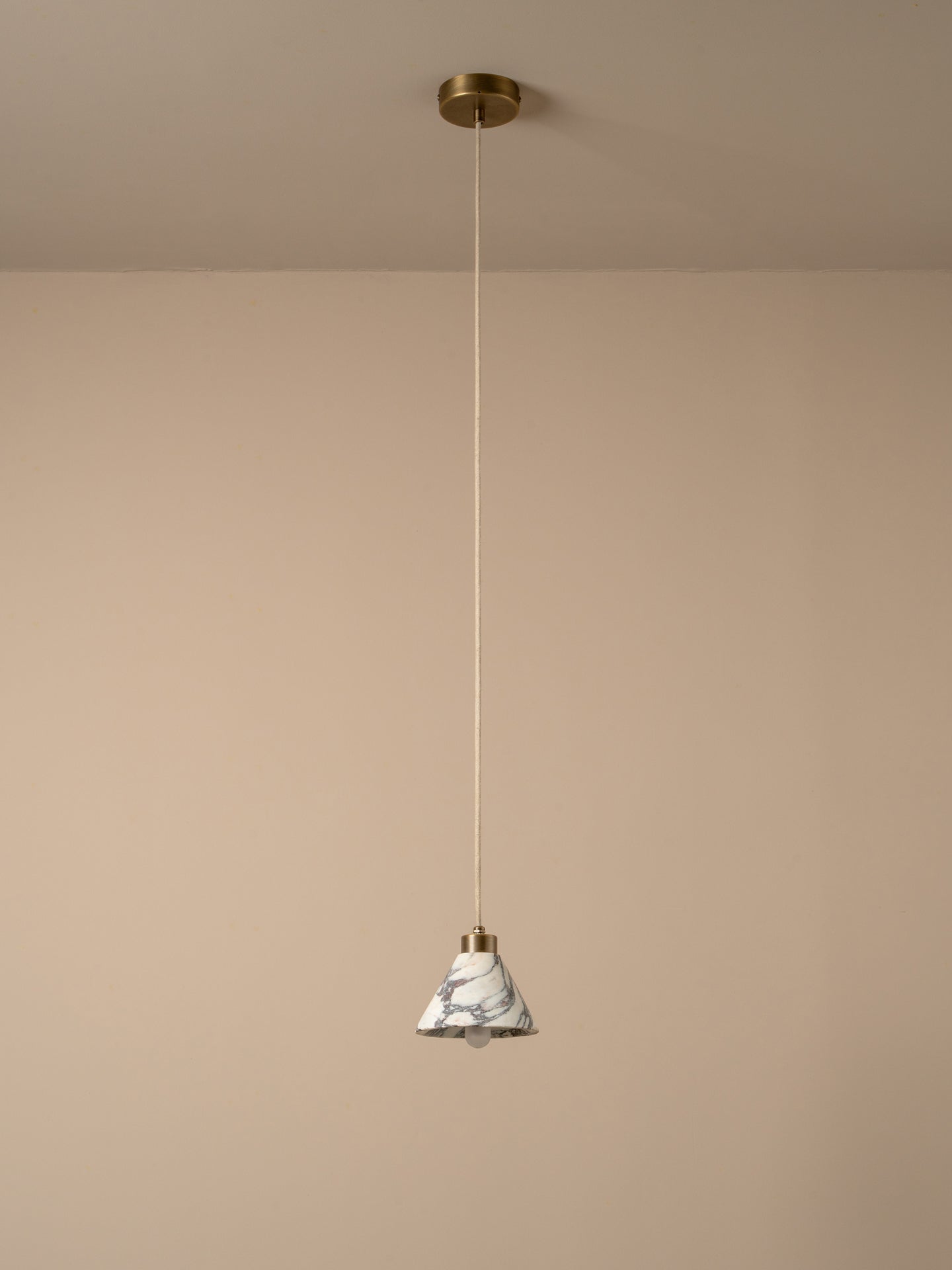 A modern pendant light with a white and gray marble cone shade, suspended by a thin cord from a brass ceiling canopy against a warm beige wall.