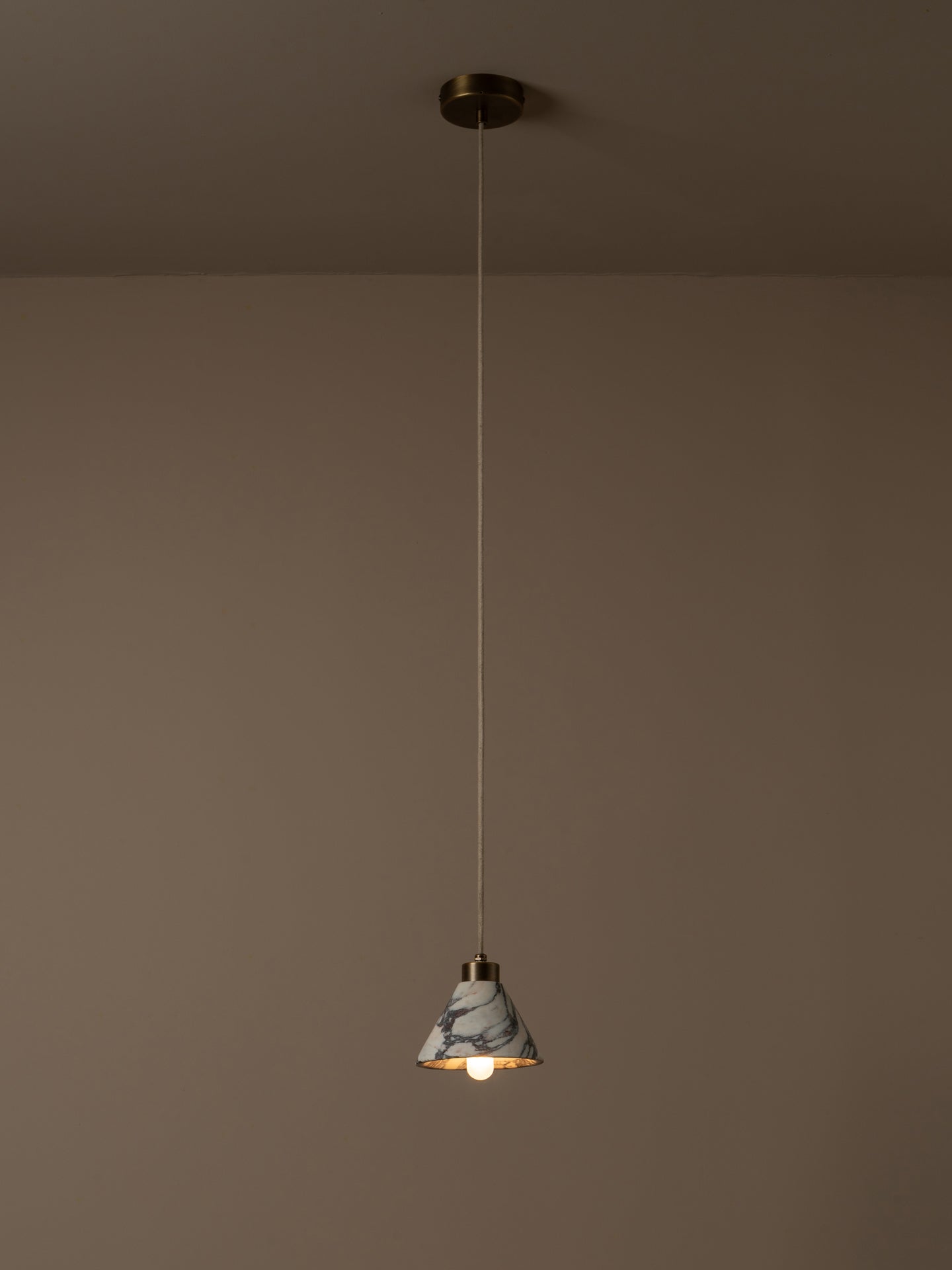 A single pendant light with a white and gray marble-patterned shade and a warm brass finish, suspended by a thin cord from a matching ceiling canopy against a warm taupe wall.