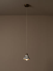 A single pendant light with a white and gray marble-patterned shade and a warm brass finish, suspended by a thin cord from a matching ceiling canopy against a warm taupe wall.