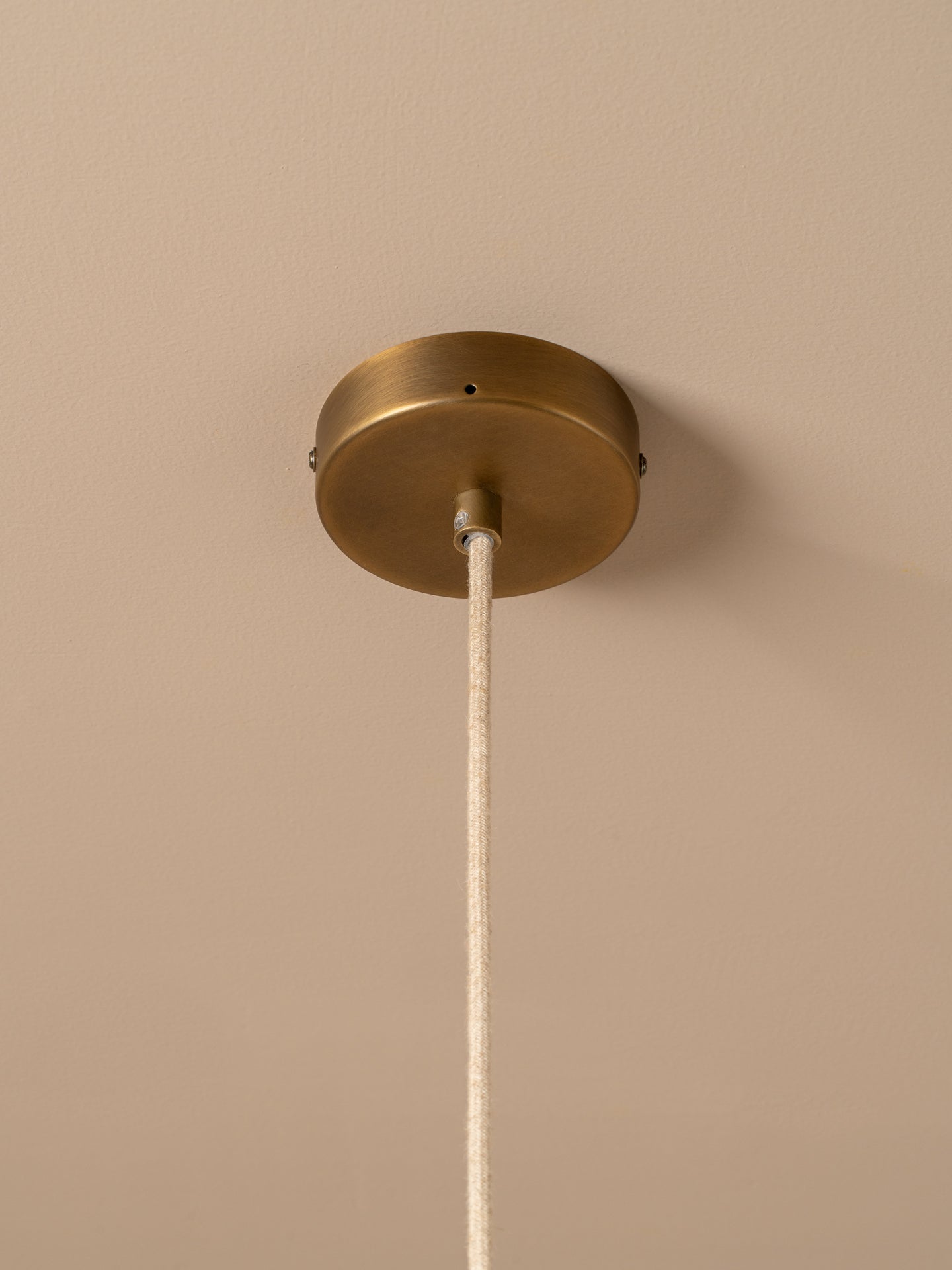 A close-up of a ceiling light fixture showing a round brass canopy mounted on a beige wall or ceiling, with a white fabric cord hanging down.