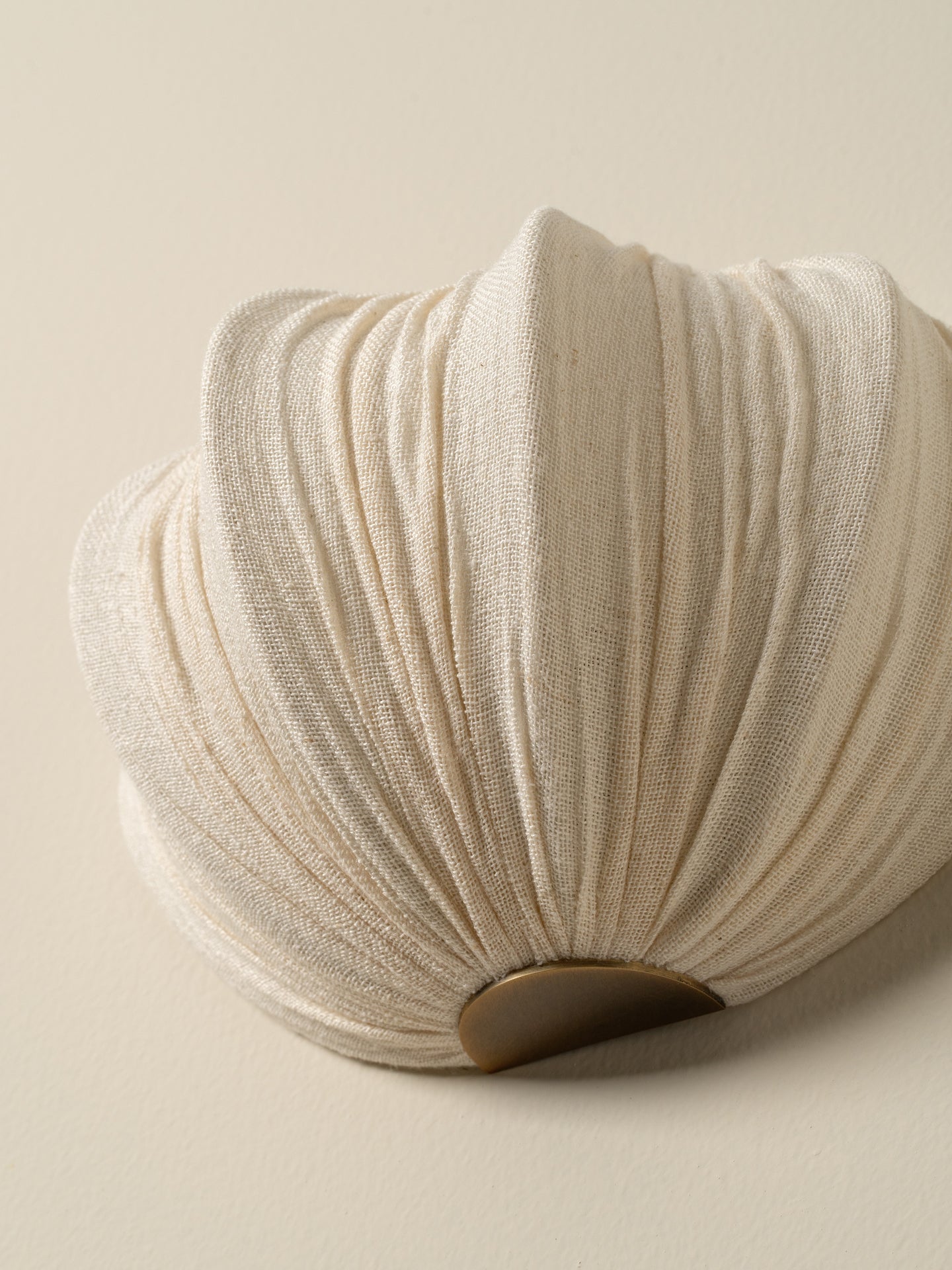 A close-up of a pleated, shell-shaped decorative object in a warm off-white or cream fabric, with a small dark brown circular accent at the base, set against a solid light beige background.