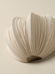 A close-up of a pleated, shell-shaped decorative object in a warm off-white or cream fabric, with a small dark brown circular accent at the base, set against a solid light beige background.