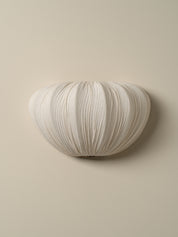 A modern wall-mounted light fixture with a pleated, ribbed fabric or paper shell in an off-white color, mounted on a solid beige wall.