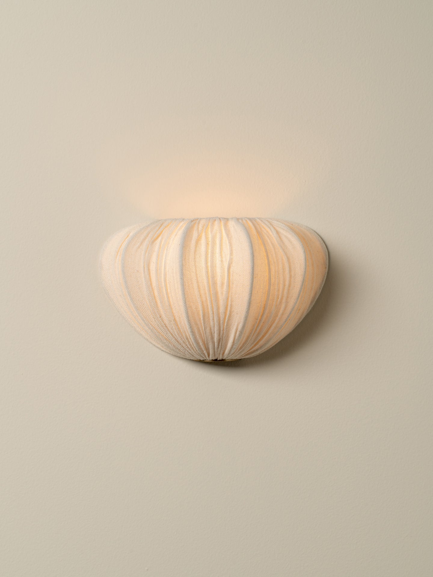 A modern wall-mounted sconce with a ribbed, organic shell-like shade in warm cream and beige tones, emitting a soft warm glow against a neutral beige wall.