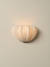 A modern wall-mounted sconce with a ribbed, organic shell-like shade in warm cream and beige tones, emitting a soft warm glow against a neutral beige wall.
