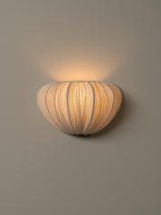 A modern wall sconce with a ribbed, organic shell-like shade, illuminated with a warm yellow glow against a neutral gray wall.