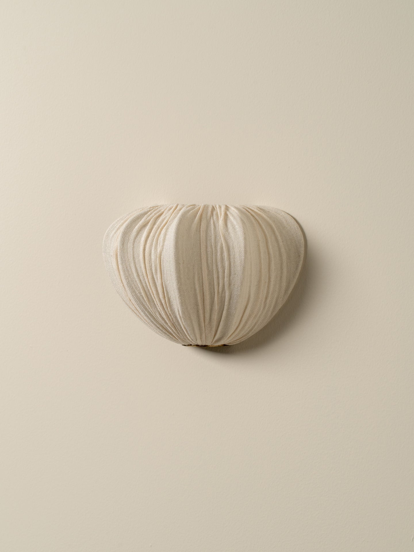 A sculptural, heart-shaped wall sconce with a pleated fabric texture in a warm cream or off-white color, mounted on a solid beige wall.