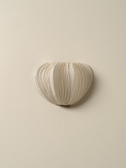 A sculptural, heart-shaped wall sconce with a pleated fabric texture in a warm cream or off-white color, mounted on a solid beige wall.