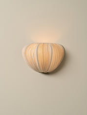 A sculptural wall sconce with a ribbed, pumpkin-like organic shape in warm beige and cream tones, emitting a soft warm glow against a plain beige wall.