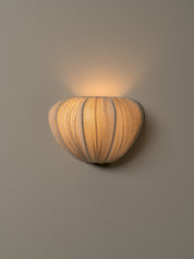 A sculptural wall sconce with a ribbed, pumpkin-like shade in warm beige and brown tones, emitting a soft warm glow against a neutral gray wall.