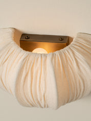 A close-up of a wall-mounted sconce with a pleated, textured cream fabric shade and a rectangular bronze metal backplate, illuminated with a warm light.