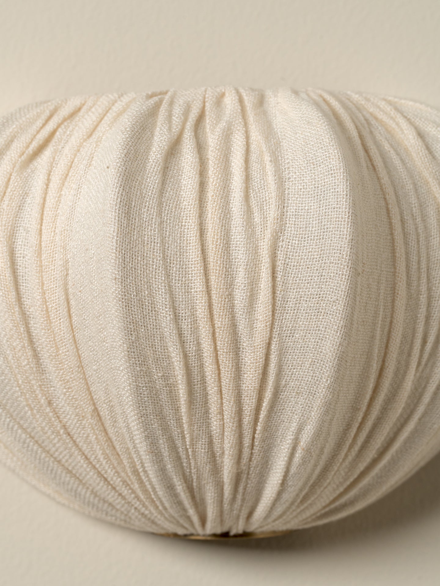 A close-up of a pleated, textured fabric cushion in a warm cream or off-white color, set against a matching neutral background.