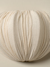 A close-up of a pleated, textured fabric cushion in a warm cream or off-white color, set against a matching neutral background.