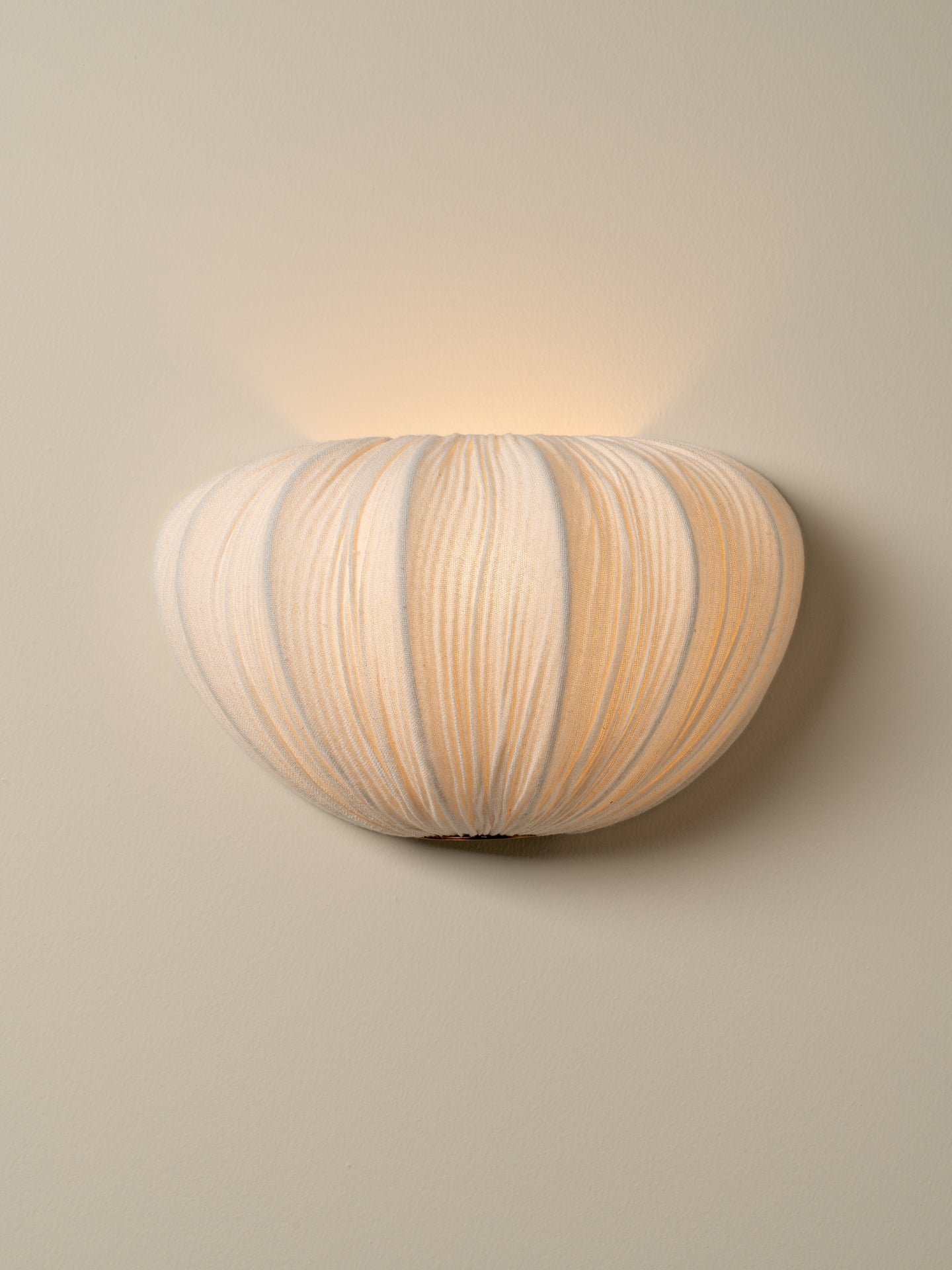 A modern wall-mounted sconce with a ribbed, pumpkin-like shade in warm off-white and beige tones, emitting a soft warm glow against a neutral beige wall.