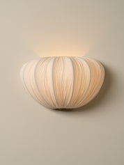 A modern wall-mounted sconce with a ribbed, pumpkin-like shade in warm off-white and beige tones, emitting a soft warm glow against a neutral beige wall.