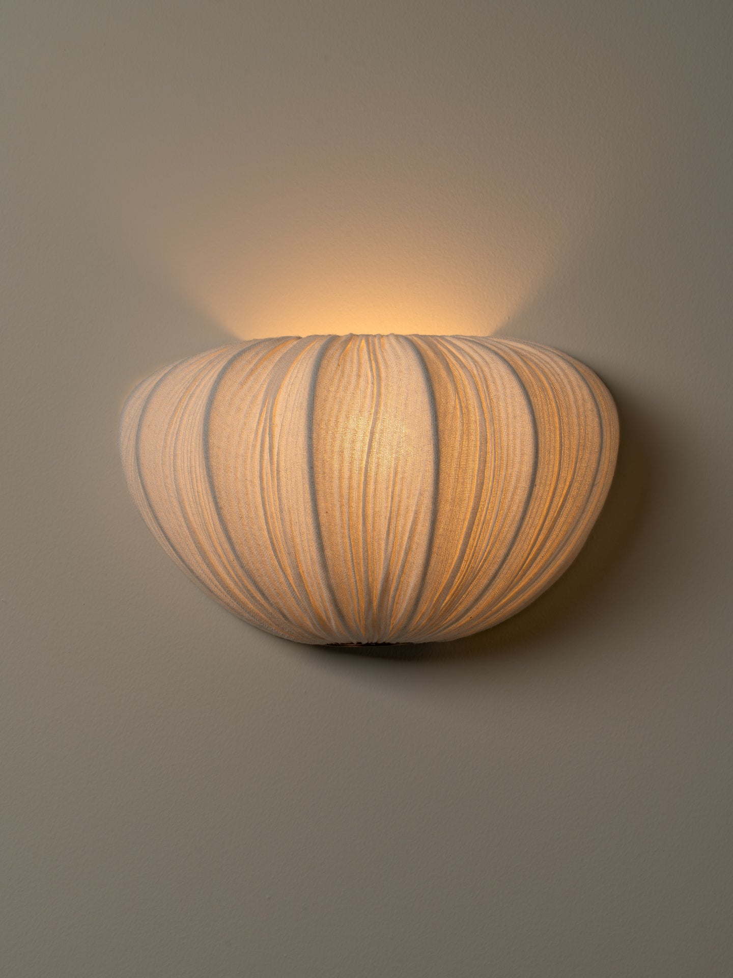 A modern wall-mounted sconce with a ribbed, pumpkin-like shade, glowing with a warm light against a neutral gray wall.