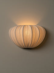 A modern wall-mounted sconce with a ribbed, pumpkin-like shade, glowing with a warm light against a neutral gray wall.