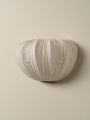 An off-white pleated fabric wall sconce mounted on a solid beige wall.